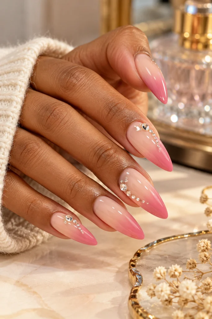 Acrylic nail designs featuring long almond extensions, a nude-to-pink ombré fade, glossy finish and delicate rhinestone accents.